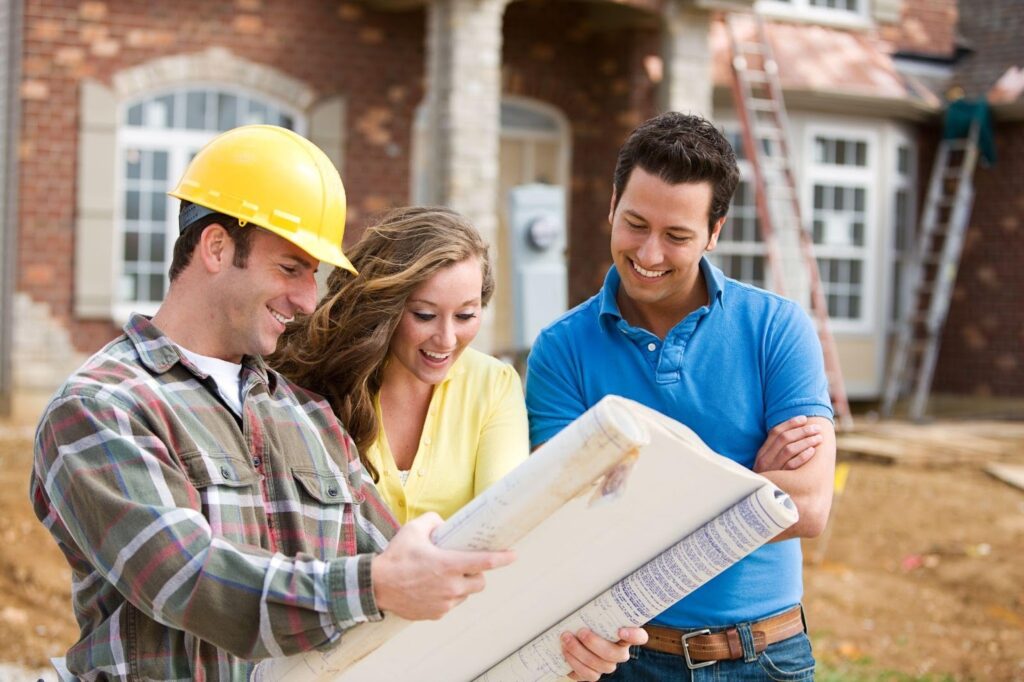 Three people standing in front of a house, examining a construction plan.