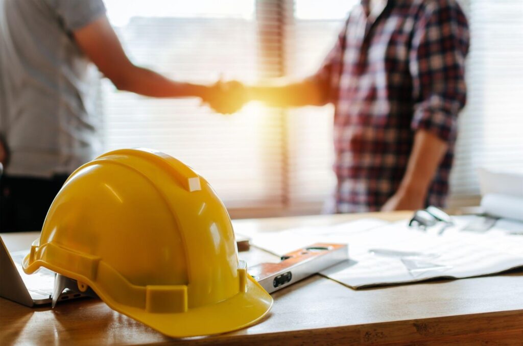 Two men shaking hands at a desk, one wearing a hard hat