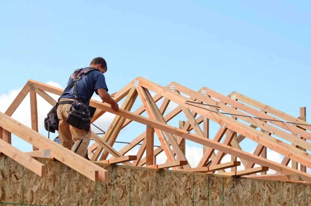 A construction worker in a safety harness working on a wooden roof frame under a clear blue sky with clouds.