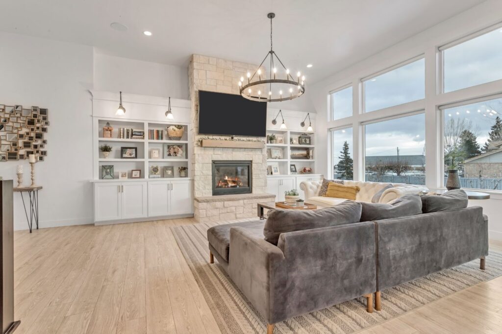 A modern family room with a stone fireplace, large windows, a grey sectional sofa, built-in white shelves, a TV, and a chandelier, featuring hardwood floors and cozy decor.